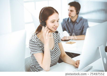 Casual dressed young woman using headset and computer while talking with customers online. Group of operators at work. Call center, business concept Casual dressed young woman using headset and computer while talking with customers online. Group of operators at work. Call center, business concept 75497552
