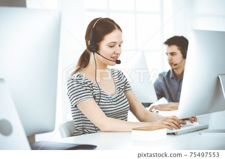Casual dressed young woman using headset and computer while talking with customers online. Group of operators at work. Call center, business concept Casual dressed young woman using headset and computer while talking with customers online. Group of operators at work. Call center, business concept 75497553