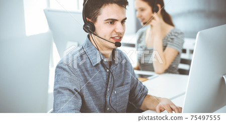 Casual dressed young man using headset and computer while talking with customers online. Group of operators at work. Call center Casual dressed young man using headset and computer while talking with customers online. Group of operators at work. Call center 75497555