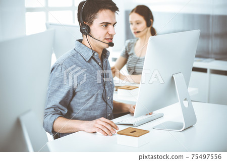 Casual dressed young man using headset and computer while talking with customers online. Group of operators at work. Call center 75497556