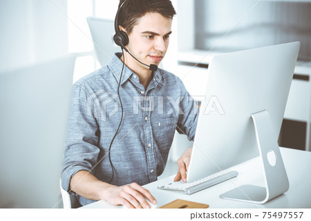 Casual dressed young man using headset and computer while talking with customers online. Call center, business concept Casual dressed young man using headset and computer while talking with customers online. Call center, business concept 75497557