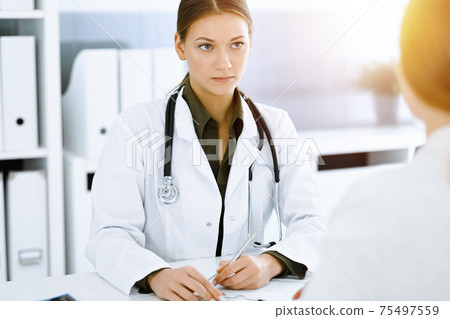 Woman-doctor and patient sitting and talking at sunny hospital office. Green color blouse suits to therapist. Medicine and healthcare concept Woman-doctor and patient sitting and talking at sunny hospital office. Green color blouse suits to therapist. Medicine and healthcare concept 75497559