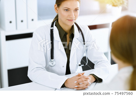 Woman-doctor and patient sitting and talking at sunny hospital office. Green color blouse suits to therapist. Medicine and healthcare concept 75497560