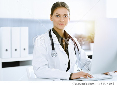 Woman-doctor typing on pc computer while sitting in sunny hospital office. Data in medicine and healthcare Woman-doctor typing on pc computer while sitting in sunny hospital office. Data in medicine and healthcare 75497561