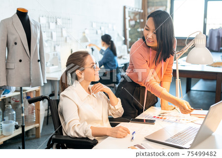 Happy young female designer in wheelchair looking at her Asian colleague pointing at laptop display 75499482