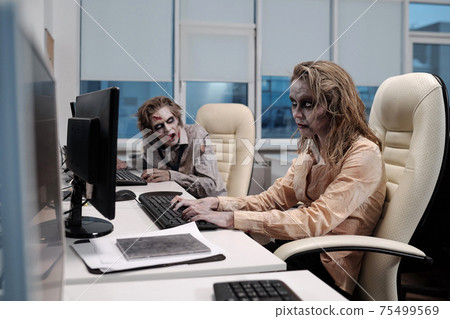 Young spooky zombie businesswoman sitting by desk in front of computer monitor and networking Young spooky zombie businesswoman sitting by desk in front of computer monitor and networking 75499569