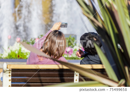 Men and women taking selfies on a park bench with a fountain Men and women taking selfies on a park bench with a fountain 75499658