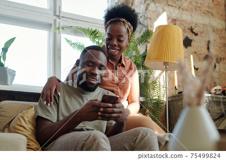 Joyful young African couple in casualwear looking at screen of smartphone held by smiling man 75499824