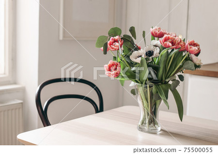 Spring flowers in glass vase on wooden table. Blurred kitchen background with old chair. Bouquet of red tulips, white anemone flowers and eucalyptus branches. Contemporary elegant, Scandinavian 75500055