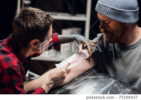 A tattoo artist bandages the young man's hand with a thin layer of petroleum jelly after applying the tattoo. A tattoo artist bandages the young man's hand with a thin layer of petroleum jelly after applying the tattoo. 75500437