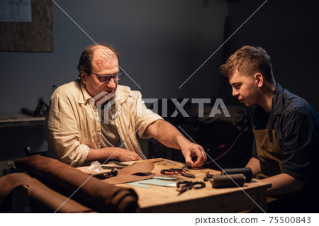 a cute young guy wants to make a gift with his own hands, an experienced shoemaker helps him make shoes out of leather. a cute young guy wants to make a gift with his own hands, an experienced shoemaker helps him make shoes out of leather. 75500843