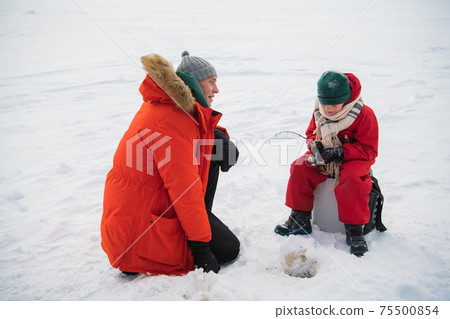 An experienced fisherman father teaches his young son to handle gear for winter fishing, they made a hole and fish in it. An experienced fisherman father teaches his young son to handle gear for winter fishing, they made a hole and fish in it. 75500854