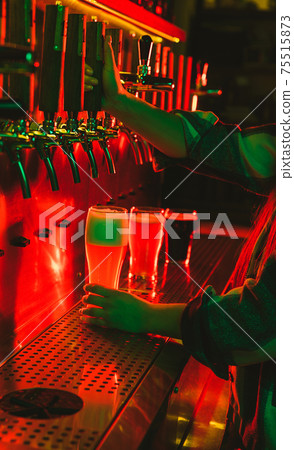 Hands of female barman pouring a cold lager beer from tap to glass in neon light Hands of female barman pouring a cold lager beer from tap to glass in neon light 75515873
