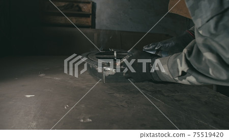 Forge workshop. Smithy. Worker in a welding hood helmet welds a part by electric welding. Sparks are reflected in the protective screen. 75519420