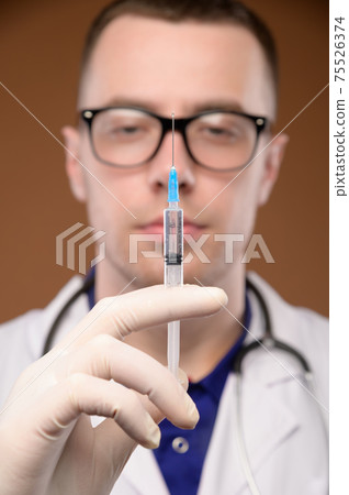 A young male Caucasian doctor holds a syringe with an injection in a gloved hand. Vaccines and vaccinations and injections concept. Focus on the syringe A young male Caucasian doctor holds a syringe with an injection in a gloved hand. Vaccines and vaccinations and injections concept. Focus on the syringe 75526374