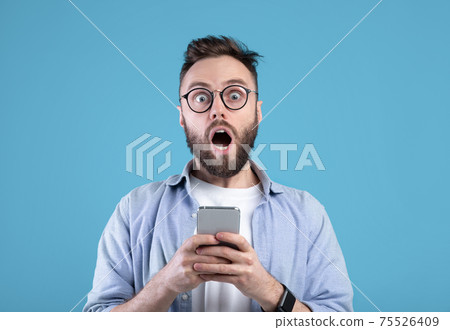 Shocked bearded guy staring at mobile phone in disbelief over blue studio background Shocked bearded guy staring at mobile phone in disbelief over blue studio background 75526409