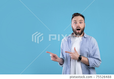 Shocked millennial man pointing at empty space in disbelief, standing with open mouth over blue background, free space 75526488