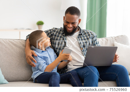 African American Father And Son Using Smartphone And Laptop Indoors African American Father And Son Using Smartphone And Laptop Indoors 75526960
