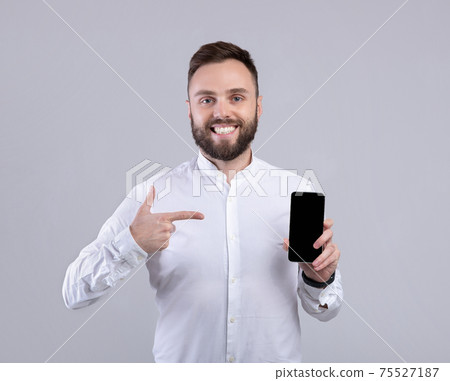 Happy bearded guy in formal wear pointing at smartphone with blank screen over grey studio background, free space Happy bearded guy in formal wear pointing at smartphone with blank screen over grey studio background, free space 75527187
