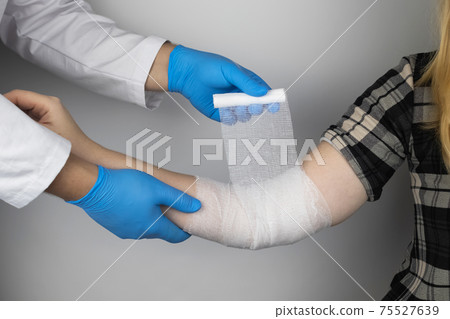 The traumatologist puts a bandage on the elbow of the female patient. The concept of help with fractures and sprains. The technique of applying a cruciform bandage. 75527639