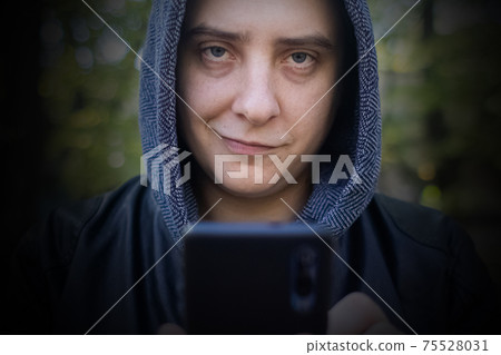 Concept. A man in a leather jacket with a hood looks at the phone and smiles like a maniac or a telephone scammer. Theft of information from a smartphone. Correspondence of a maniac on social networks Concept. A man in a leather jacket with a hood looks at the phone and smiles like a maniac or a telephone scammer. Theft of information from a smartphone. Correspondence of a maniac on social networks 75528031
