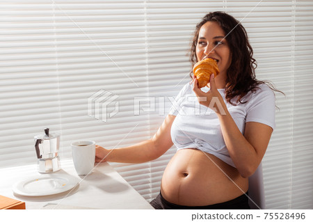 Happy pregnant young beautiful woman eating croissant and looking througt blinds during morning breakfast. Concept of pleasant morning and positive attitude during pregnancy 75528496