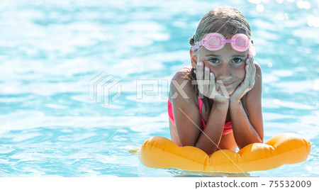Young girl rests on floating pillow inside the pool with swimming goggles on her forehead, smiling Young girl rests on floating pillow inside the pool with swimming goggles on her forehead, smiling 75532009