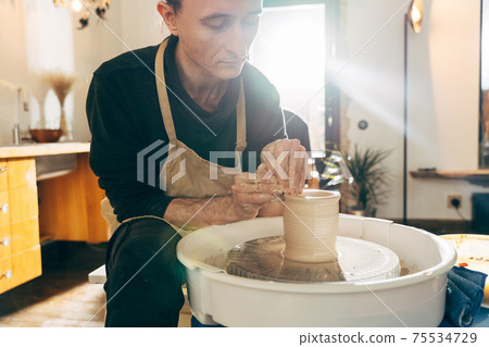 Master sitting at the potter's wheel and holding unfired clay cup Master sitting at the potter's wheel and holding unfired clay cup 75534729