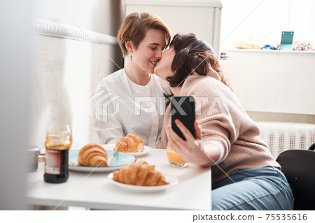 Woman holding smartphone and kissing her girlfriend while making selfie together 75535616