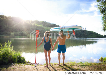 Rear view of senior couple carrying paddleboard by lake in summer. Rear view of senior couple carrying paddleboard by lake in summer. 75536780