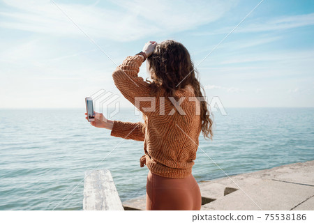 Female from the back filming video or making a picture on the beach using her smartphone Female from the back filming video or making a picture on the beach using her smartphone 75538186
