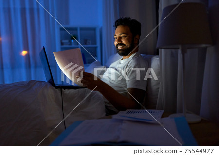 indian man with laptop and papers in bed at night indian man with laptop and papers in bed at night 75544977