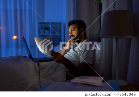indian man with laptop and papers in bed at night 75545049