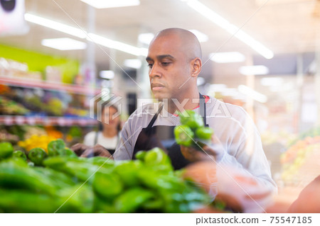 Merchandiser in an apron lays out ripe bell peppers on supermarket counter Merchandiser in an apron lays out ripe bell peppers on supermarket counter 75547185