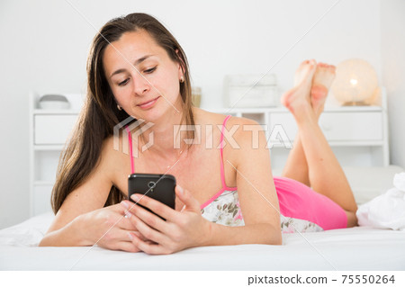 Adult positive woman using her phone while lying in bed Adult positive woman using her phone while lying in bed 75550264