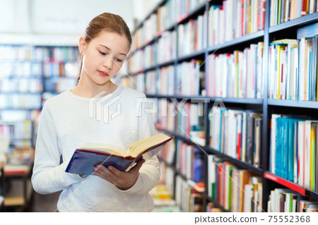 Intelligent preteen girl standing alone near bookcase in library 75552368