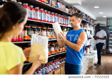 Focused muscular man looking for necessary sport nutrition products in shop, reading content label 75553505