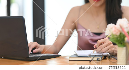 Cropped image woman is taking notes and using a computer laptop at the wooden working desk. Cropped image woman is taking notes and using a computer laptop at the wooden working desk. 75558221
