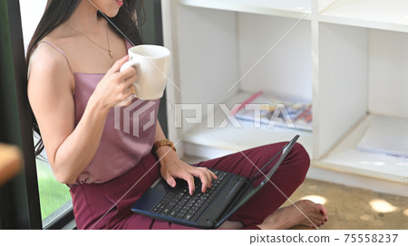 Cropped image woman is drinking coffee and using a computer laptop while sitting in the living room. Cropped image woman is drinking coffee and using a computer laptop while sitting in the living room. 75558237