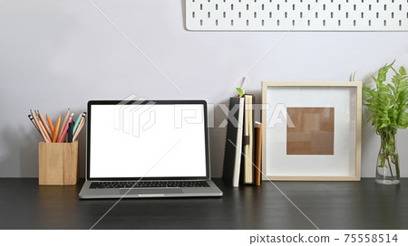 Laptop on working desk along with white blank screen computer, books, notebook, pencil holder, picture frame, potted plant putting together on it with white cement wall as background. Laptop on working desk along with white blank screen computer, books, notebook, pencil holder, picture frame, potted plant putting together on it with white cement wall as background. 75558514