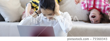 Woman sits on floor next to the sofa with laptop covering her ears with crabs from behind the children indulge 75560343