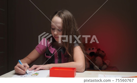 The girl draws a family on a white sheet of paper with a bright colored felt-tip pen. The child is engaged in creative work while sitting at the table in the children's room at night. Spend the The girl draws a family on a white sheet of paper with a bright colored felt-tip pen. The child is engaged in creative work while sitting at the table in the children's room at night. Spend the 75560767