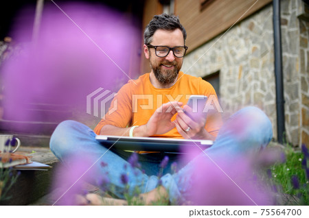 Mature man with smartphone working outdoors in garden, green home office concept. 75564700
