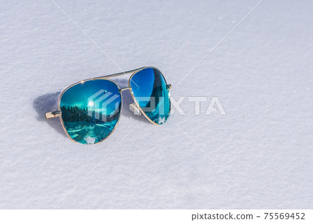 Sunglasses with blue lenses in which the sun and the forest are reflected lie on the snow Sunglasses with blue lenses in which the sun and the forest are reflected lie on the snow 75569452