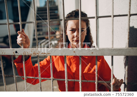 Young woman in orange suit behind jail bars. Female in colorful overalls portrait. Law and justice concept. Young woman in orange suit behind jail bars. Female in colorful overalls portrait. Law and justice concept. 75572360