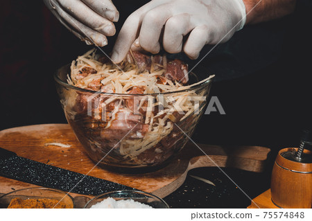A large piece of pork is cut into pieces, marinated by the chef with spices, and the chef rubs the potatoes on a grater to the marinade. A large piece of pork is cut into pieces, marinated by the chef with spices, and the chef rubs the potatoes on a grater to the marinade. 75574848