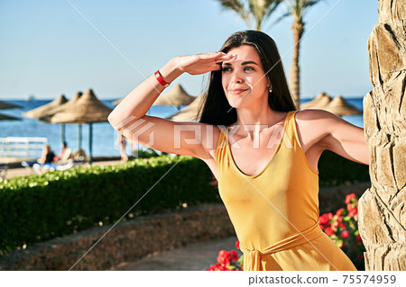 Beautiful caucasian lady with dark hair in yellow dress stands by a palm tree at luxury tropical resort seaside hotel shielding eyes with hand Beautiful caucasian lady with dark hair in yellow dress stands by a palm tree at luxury tropical resort seaside hotel shielding eyes with hand 75574959