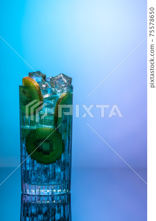 A glass of water with ice and slices of kiwi standing on a mirror table in neon light. Body detoxification or detoxication concept 75578650