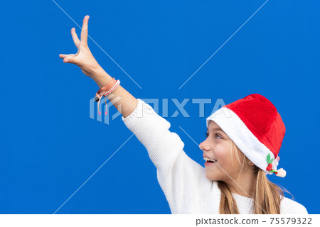 Joyful Young girl in Santa hat gesturing on blue background. 75579322