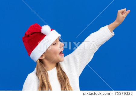 Joyful Young girl in Santa hat gesturing on blue background. 75579323